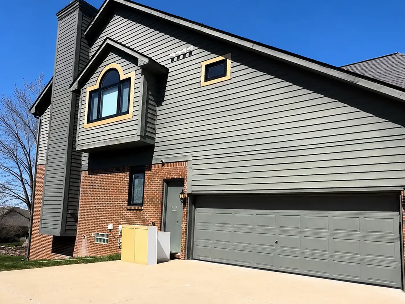 Front of a two-storey house before painting: sage-grey lap siding across the gable, a butter-yellow surround on the arched dormer window, and a grey garage door.