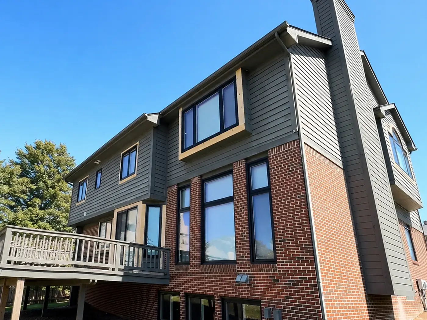 Rear of the same house before painting: grey-green siding with tan window surrounds, a weathered raised deck, and a sided chimney running the full height.