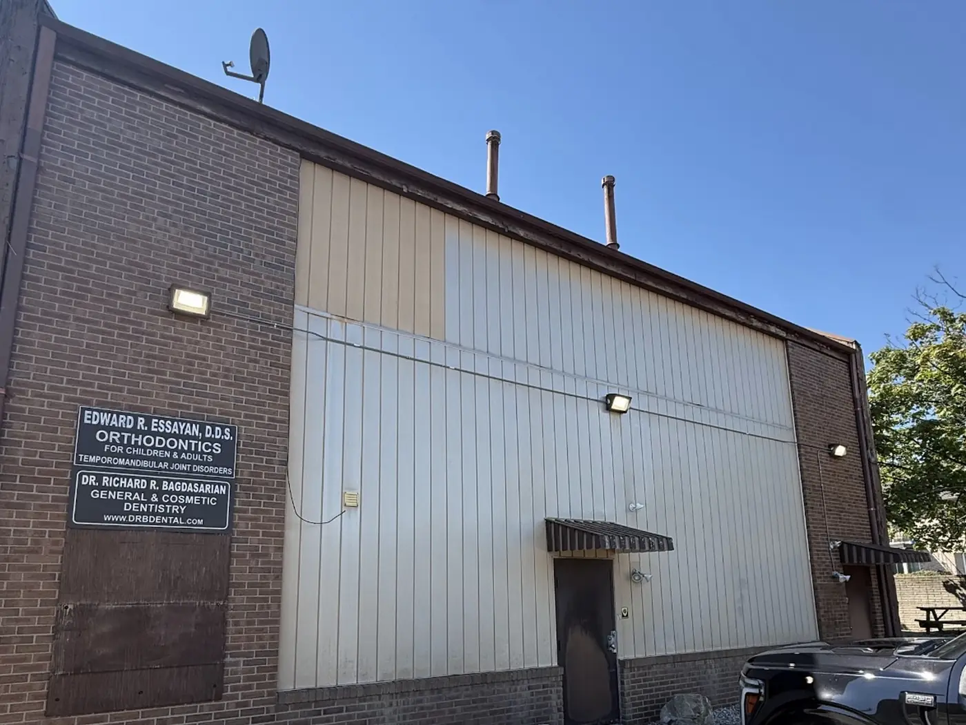Side wall of a single-storey office building before painting: metal panels weathered into mismatched bands of cream and grey, with a bare plywood panel and sun-bleached brown trim above the brick.