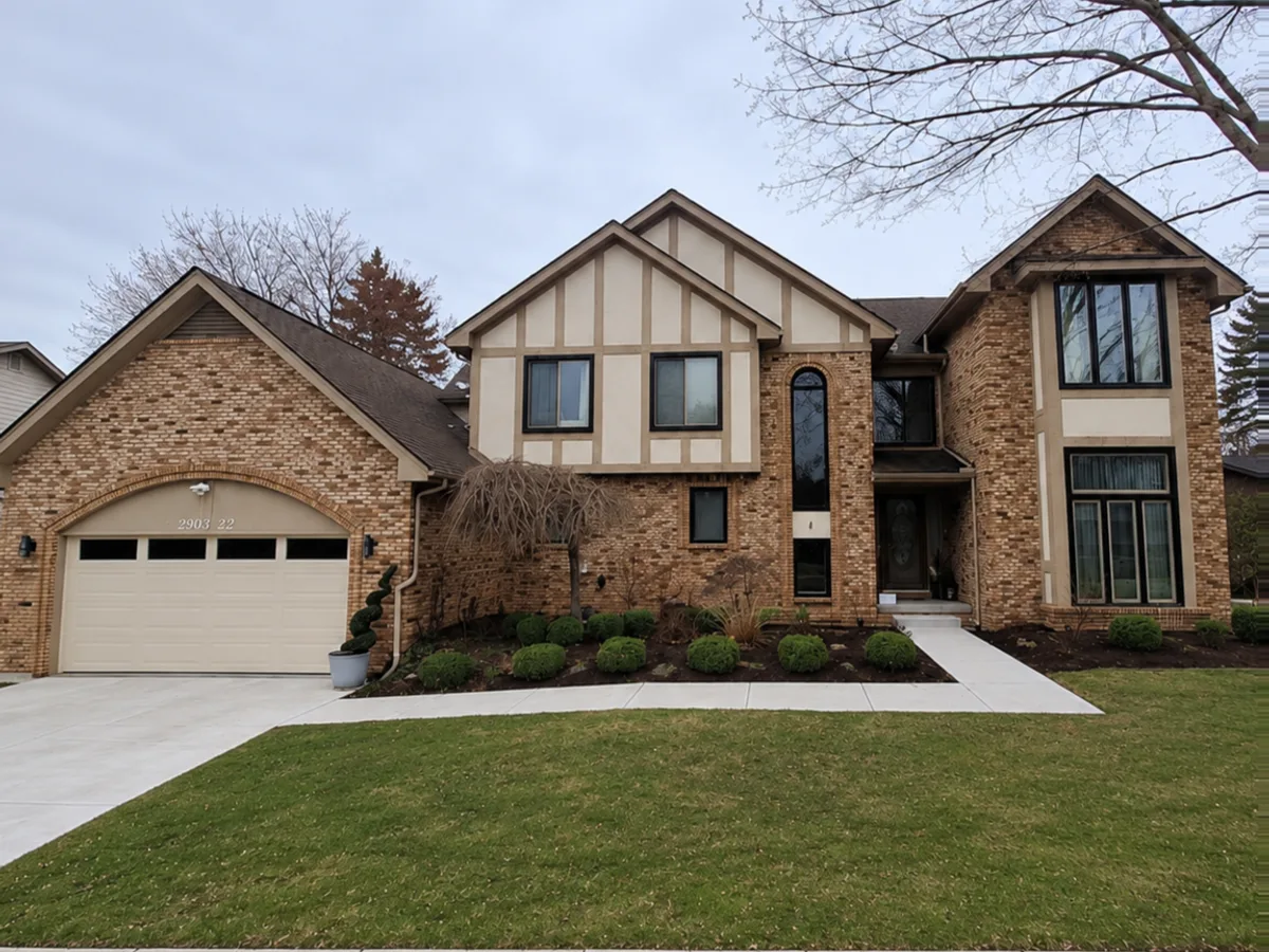 Two-storey Tudor-style house before painting: tan brick across both storeys, beige stucco and brown half-timbering in the gables, and a cream garage door.
