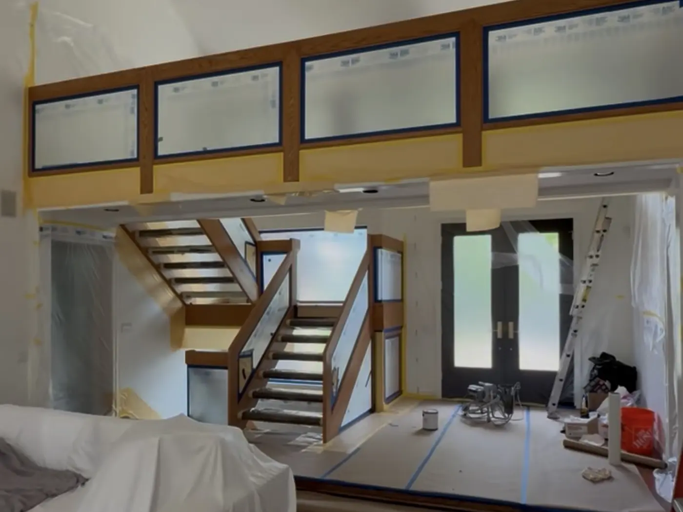 Double-height stairwell before painting: oak window frames and railings masked with blue tape and film, furniture sheeted, and drop cloths taped across the floor.